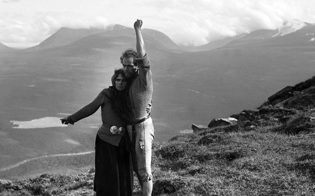 Film still from BERG-EYVIND AND HIS WIFE: A man and a woman stand in a lonely mountain landscape. They are holding each other and each has one arm stretched out from them.