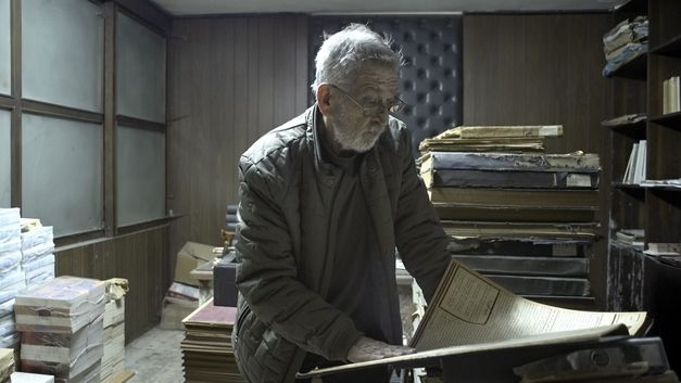 Ein älterer Mann steht zwischen Aktenstapeln und liest ein großes Dokument.
