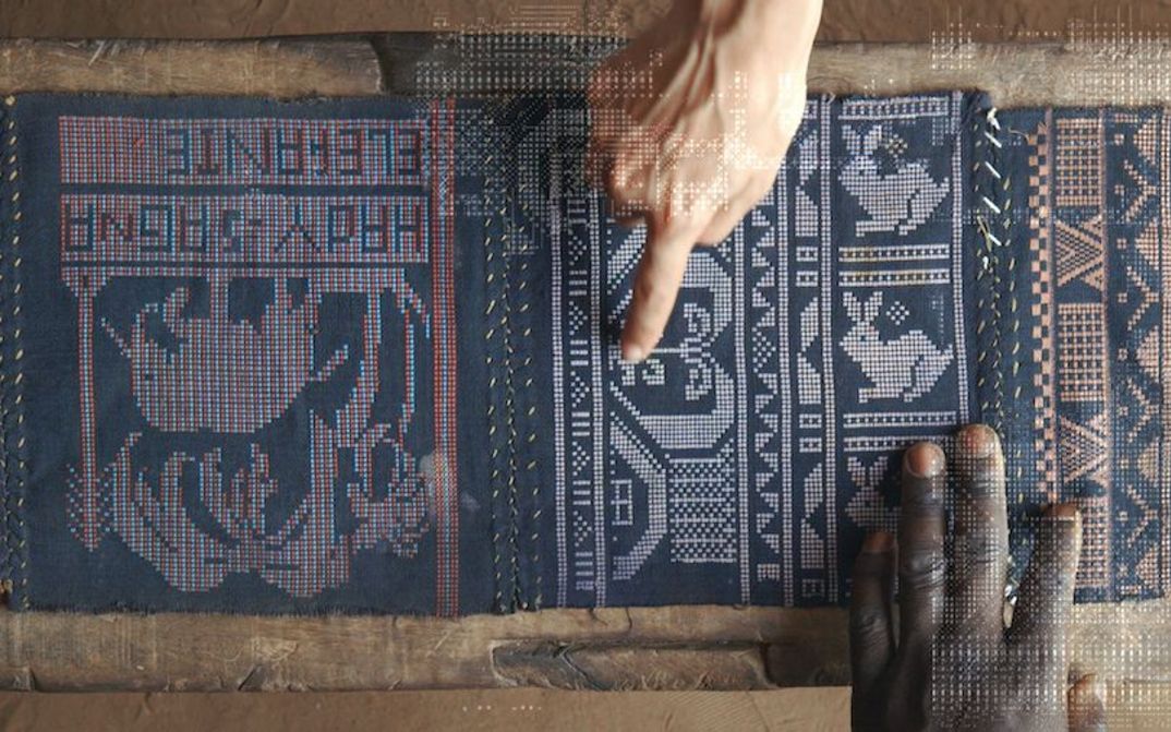 Film still from QUANTUM CREOLE: View of an elaborately woven cloth. A black and a white hand point to it.