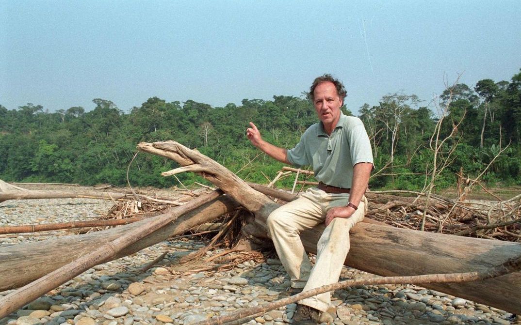 Film still from MEIN LIEBSTER FEIND: Werner Herzog sits on a lying tree trunk and points to the jungle behind him.