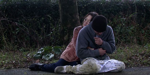 A man and a woman are sitting on the ground on a park path. She sits behind him and hugs him. 