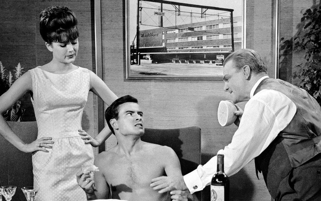 Film still from ONE, TWO, THREE: A young man with a naked upper body is sitting at a table eating. To his left and right are a woman and a man who are looking at him disapprovingly.
