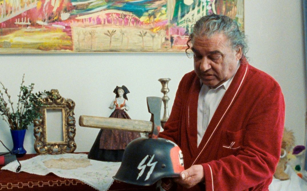 Filmstill from WANKOSTÄTTN: A man with grey hair and a red bathrobe is holding a helmet with SS lettering on it. There is an axe in the helmet and red paint around it.