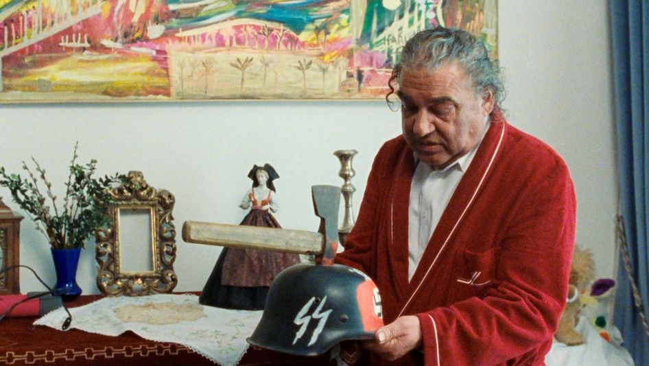 Filmstill from WANKOSTÄTTN: A man with grey hair and a red bathrobe is holding a helmet with SS lettering on it. There is an axe in the helmet and red paint around it.