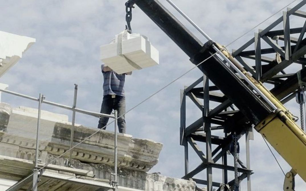 Still from the film „Onun Haricinde, yiyim“ by Eren Aksu. A construction site for a building with a crane. A man on the construction site receives building elements with the crane