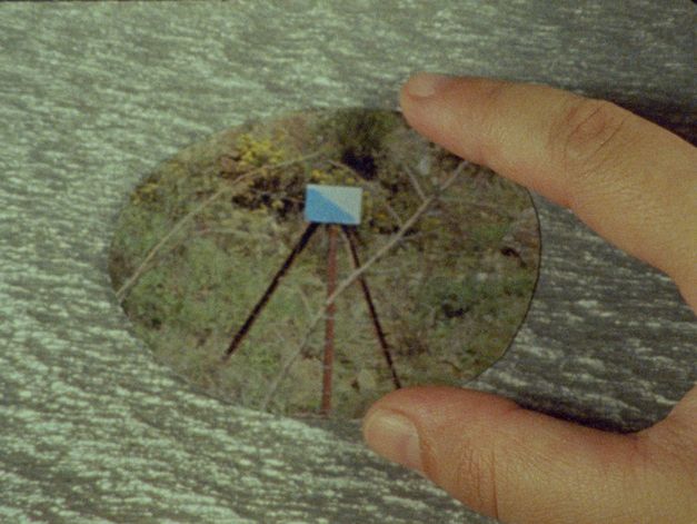 Filmstill aus dem Film „Portales“ von Elena Duque. Eine in Großaufnahme gefilmte Hand legt ein Foto, das einen blauen Bilderrahmen in einem Feld zeigt, auf ein Foto, das das glitzernde Wasser eines Flusses in Großaufnahme zeigt.