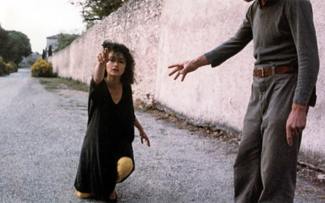 Film still from NEUER ENGEL. WESTWÄRTS. A woman kneels on a street and points to something outside the picture.