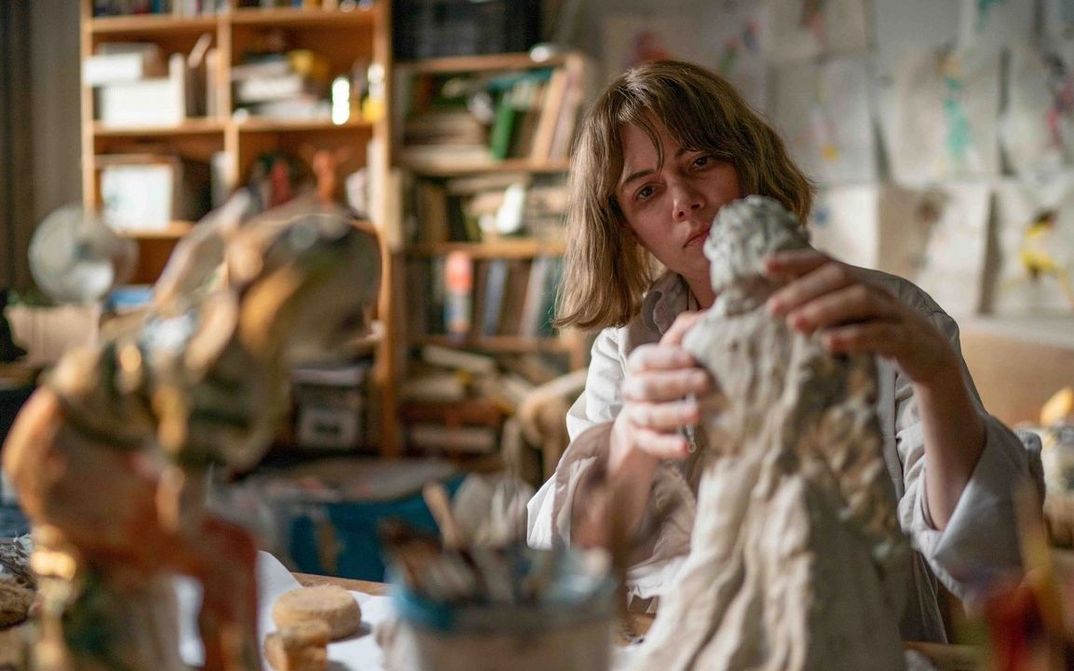Film still from SHOWING UP: A blonde woman works on a small clay sculpture. A cluttered room can be seen blurred in the background.