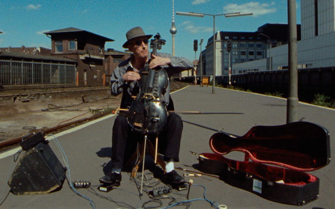 Film still from BERLIN - BAHNHOF FRIEDRICHSTRASSE: A man plays the cello and sits on a street. Next to it is an old railway station building and the Berlin television tower in the background.