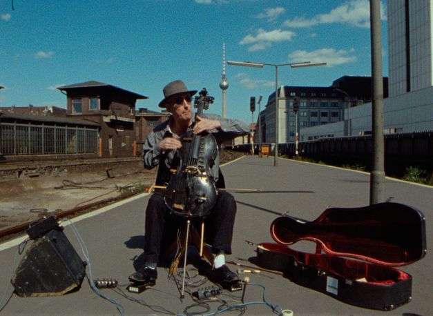 Filmstill aus BERLIN - BAHNHOF FRIEDRICHSTRASSE: Ein Mann spielt Cello und sitzt auf einer Straße. Daneben ein altes Bahnhofsgebäude und im Hintergrund der Berliner Fernsehrturm.