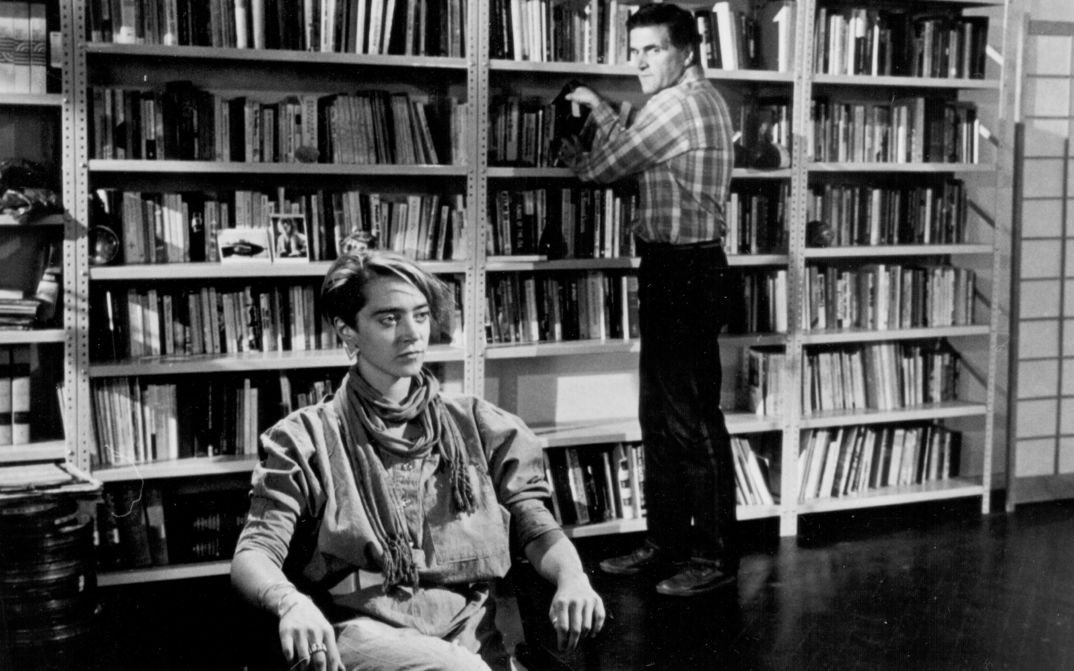 Film still from THE MAN WHO ENVIED WOMEN: A man stands at a bookshelf, a woman sits in front of it.