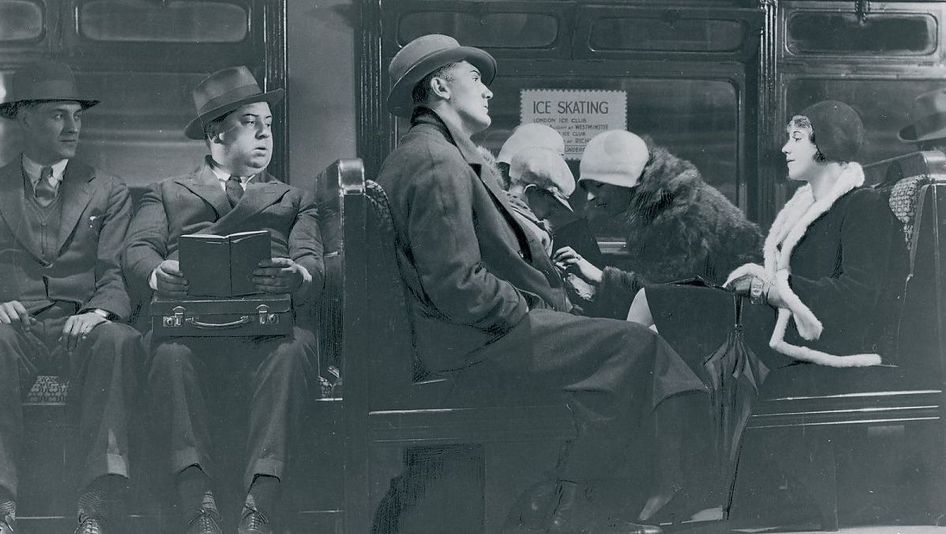 Filmstill aus BLACKMAIL: Mehrere elegant gekleidete Menschen sitzen in einem Zug. Auf einem Sitz ist Alfred Hitchcock zu sehen. Er hält ein aufgeschlagenes Buch in den Händen, blickt aber zu den anderen Passagieren.