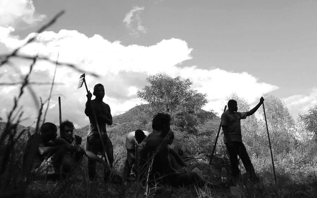 Film still from GEOLOGY OF SEPARATION: A group of men stand and sit between trees in a hilly landscape.