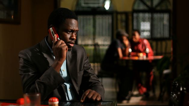 Film still from CONFUSION NA WA: A man in a suit on the phone. In the background you can see two people sitting at a table, out of focus. 