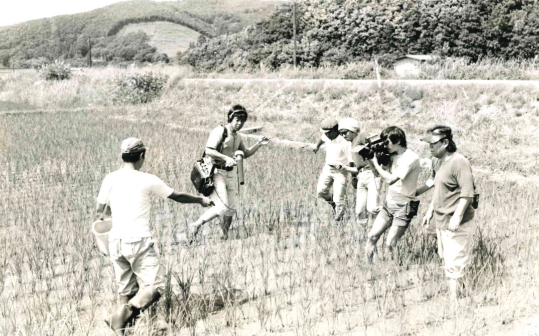 Film still from „Nippon“: Furuyashiki Village: A few people are standing in a rice field, a small film crew is recording.