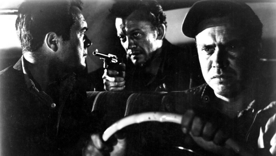 Film still from THE HITCH-HIKER: Three men are sitting in a car. The man in the back seat points a revolver at the man in the passenger seat.