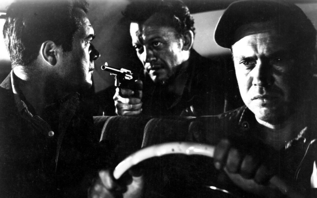 Film still from THE HITCH-HIKER: Three men are sitting in a car. The man in the back seat points a revolver at the man in the passenger seat.