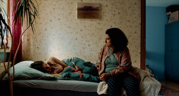 Filmstill aus WENN DU ANGST HAST NIMMST DU DEIN DEIN HERZ IN DEN MUND UND LÄCHELST von Marie-Luise Lehner. Das Bild zeigt ein buntes Schlafzimmer mit einem Bett, auf dem eine junge Person liegt. Eine Frau sitzt auf der Bettkante. 