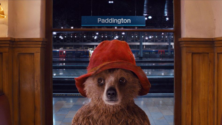 Filmstill aus PADDINGTON: Ein Teddybär mit einem roten Hut steht in einem Bahnhof. Hinter ihm sieht man die Gleise und eine Tafel mit der Aufschrift "Paddington".