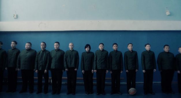 Film still from CADET by Adilkhan Yerzhanov. A group of pupils in school uniforms stand lined up in front of a wall. One of them has longer hair than the others. 