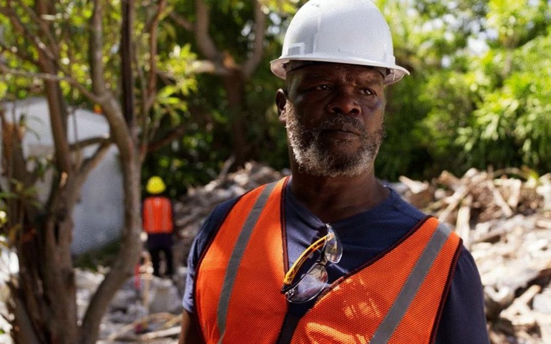 Film still from MOUNTAINS: A man wearing a safety helmet and vest stands in front of a demolished building.
