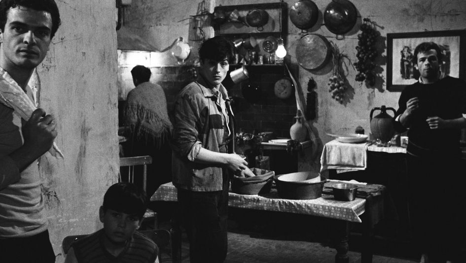 Filmstill from ROCCO E I SUOI FRATELLI: Several men and a woman in the kitchen. There are pots on the table and some kitchen utensils and decorations hanging on the wall.