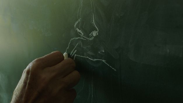 Film still from Alban Muja’s film “Mua besoj më shpëtoj portreti”. A hand in close-up draws with chalk on a blackboard