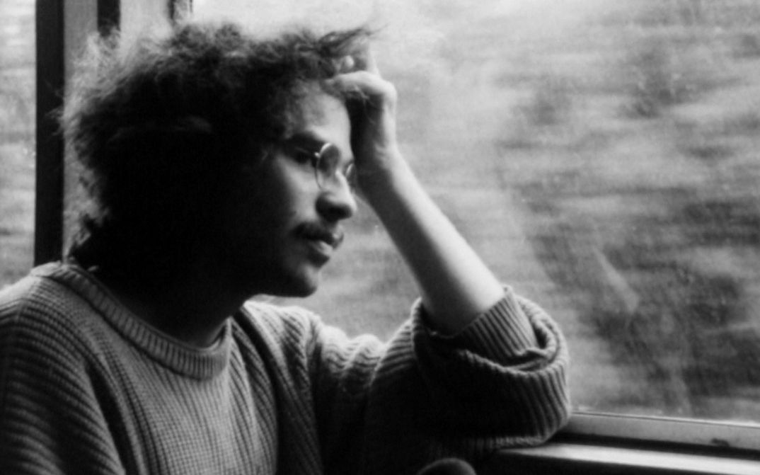Film still from IM ÜBERGANG: A man with glasses sits at the window in the train, rests his head on his hand and looks out of the window.