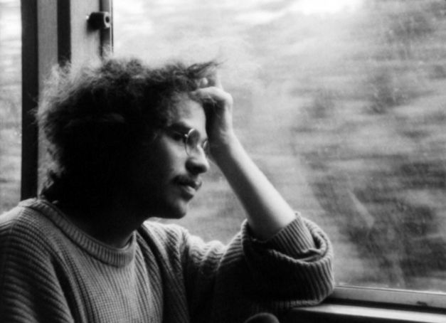 Filmstill aus IM ÜBERGANG: Ein Mann mit Brille sitzt am Fenster im Zug, stützt seinen Kopf auf der Hand ab und schaut aus dem Fenster.