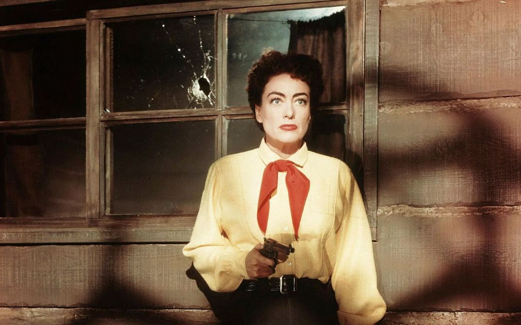 Filmstill from JOHNNY GUITAR: A woman in a yellow shirt and red scarf holds a pistol in her hand, looks startled and stands in front of a wooden house and a window with a bullet hole in it.