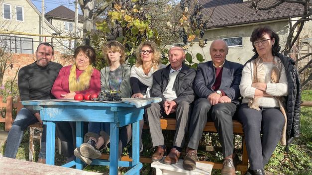 Sieben Leute sitzen nebeneinander auf einer Bank im Garten vor einem Tisch mit Obst, im Hintergrund ein Wohnhaus.