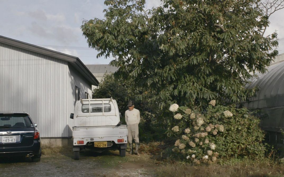 Film still from JAPAN - BIG LAGOON VILLAGE: A man in rubber boots stands between trees and a pick-up truck.
